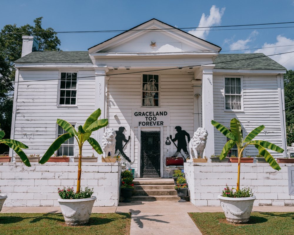Graceland Too’s white facade, with lion statues, Elvis silhouettes, banana plants, and a Graceland Too Forever sign.
