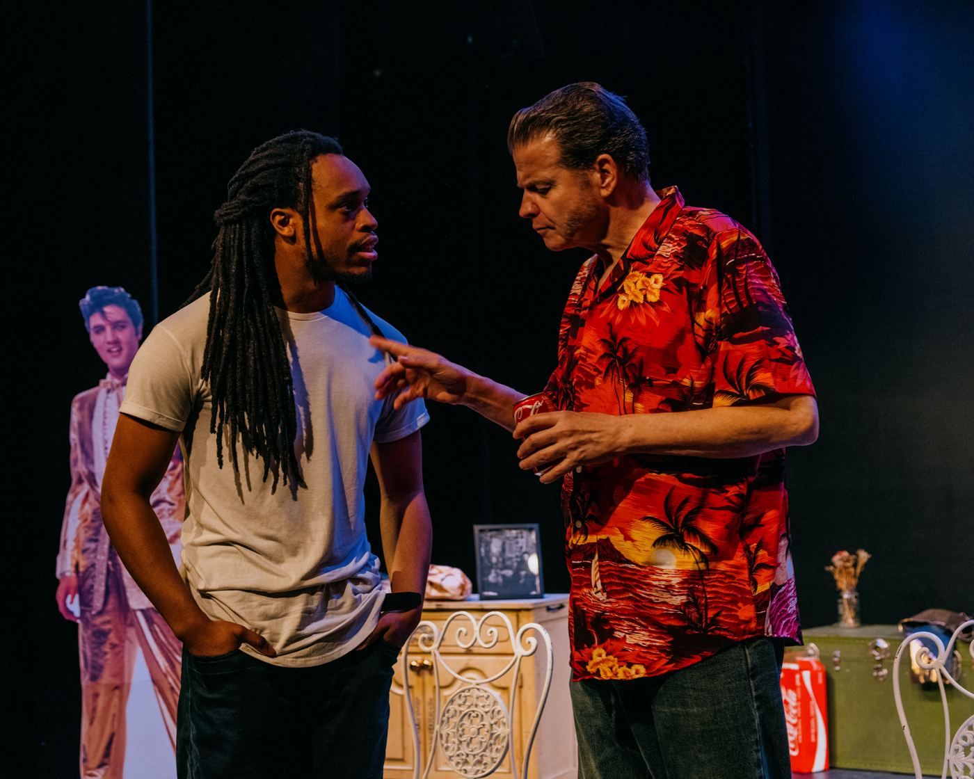 Dwight and Paul face one another on stage, both fully visible, beside a cardboard Elvis cutout.