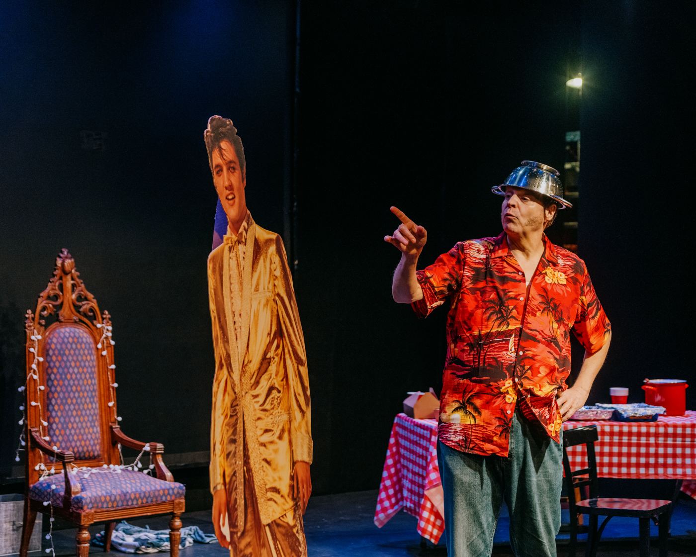 Paul wears a colander as a hat and points beside a life-size Elvis cutout, with a throne chair and red checked table behind him.